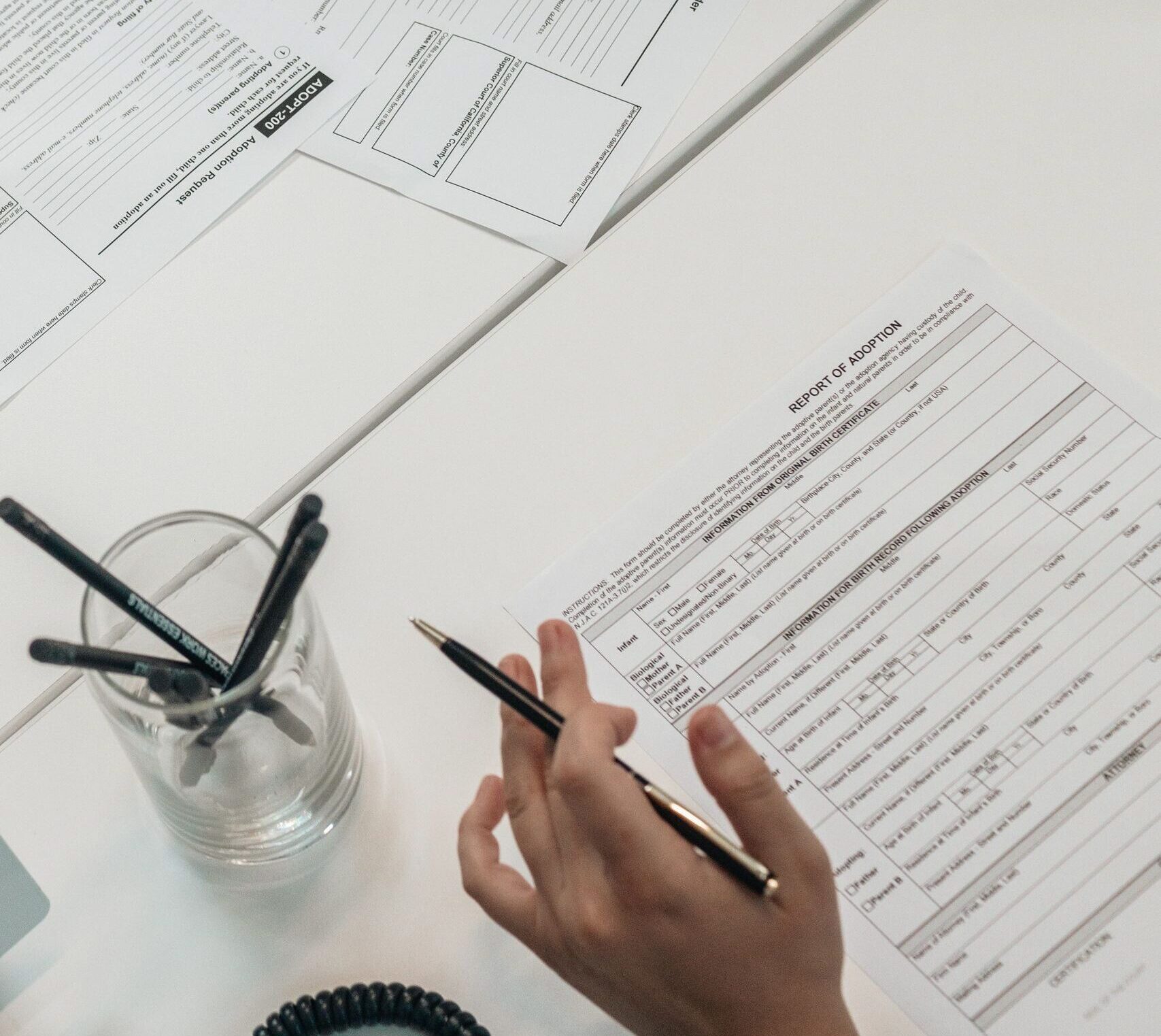 A person holds a pen while examining business documents on a white desk, top view.